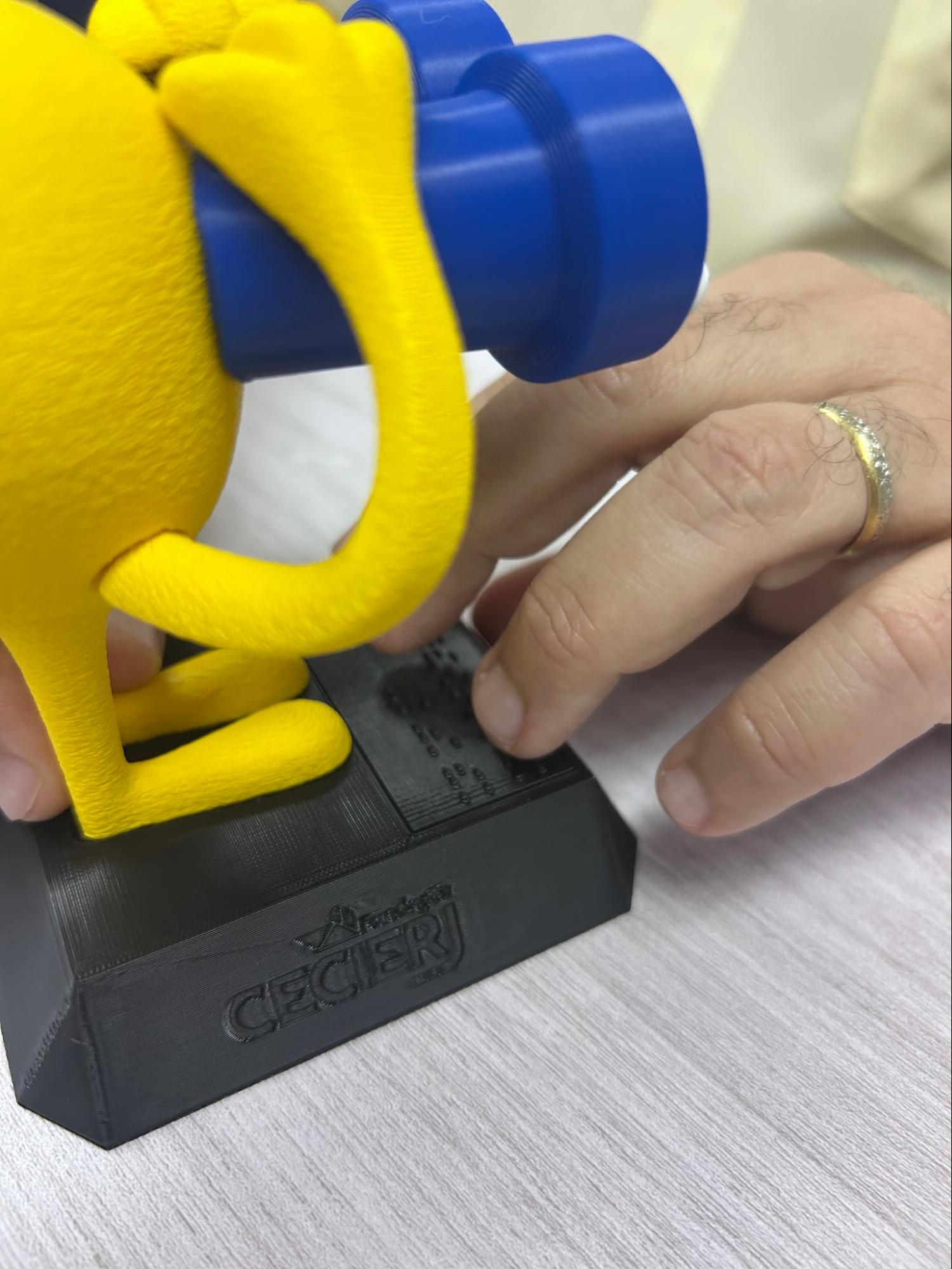 Audiodescrição: Fotografia vertical dos dedos do consultor tocando o texto em braile no pedestal do mascote. Fim da audiodescrição.<br />
