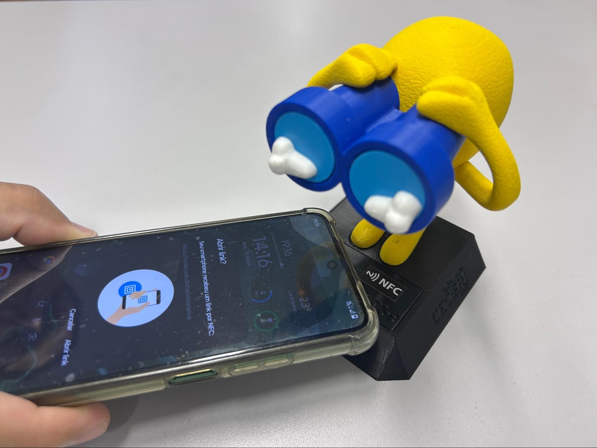 Audiodescrição: fotografia horizontal de mão segurando um celular proximo à etiqueta NFC que está no pedestal do mascote 3D. A tela do celular exibe o texto “Abrir link?”. Fim da audiodescrição.