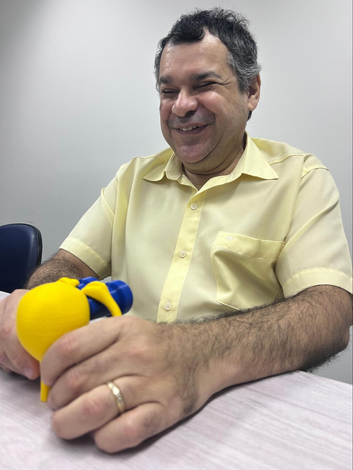 Audiodescrição: Fotografia vertical do consultor sentado com as mãos tocando o Mascote 3D sobre uma mesa. Ele é um homem branco, com cabelos grisalhos. Está sorridente. Usa camisa de manga curta amarela. Fim da audiodescrição.<br />
