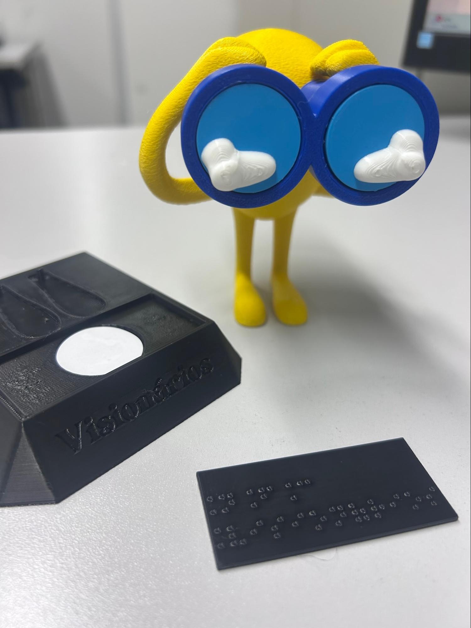Audiodescrição: Fotografia vertical do mascote 3D. Á esquerda um pedestal preto no formato de um trapézio, com uma etiqueta branca redonda no topo, e a palavra VISIONÁRIOS em baixo relevo na face frontal. Á frente do mascote uma plaquinha preta em com as palavras NFC e Visionários em braile. Fim da audiodescrição.<br />
