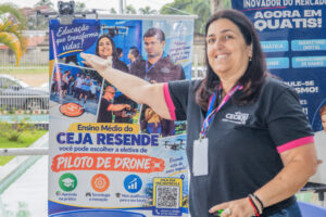 Servidora da Fundação Cecierj sorri ao lado de painel informativo do CEJA Resende que divulga a nova eletiva de Piloto de Drone, indicando um pequeno drone no topo